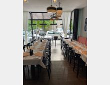 Restaurantinnenraum mit gedeckten Tischen und weißen Tischdecken. Schwarze Stühle stehen entlang der Tische, große Fensterfront mit weißen Vorhängen und Blick auf geparkte Autos und grüne Bäume im Außenbereich.