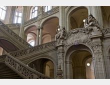 Großes historisches Treppenhaus mit aufwendig verzierten Steinbalustraden, Bögen und reich dekorierter Architektur. Figuren und ornamentale Reliefs schmücken die Wände und Balustraden, während hohe Fenster natürliches Licht einlassen.