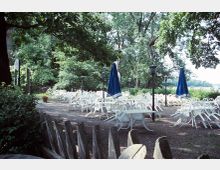 Außenterrasse eines Cafés oder Biergartens mit zahlreichen weißen Plastikstühlen und -tischen, von denen einige umgekippt sind. Umgeben von üppigem Grün und Bäumen, stehen vereinzelt blaue Sonnenschirme sowie Laternen.