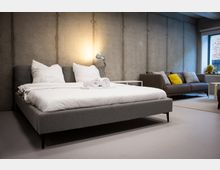 Modernes Schlafzimmer im Loft-Stil mit Betonwänden, großem grauen Polsterbett mit weißer Bettwäsche und zusammengerollten Handtüchern sowie einer Stehlampe am Nachttisch. Im Hintergrund stehen ein graues Sofa mit Kissen und ein großes Fenster, das Tageslicht in den Raum lässt.
