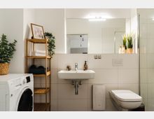Modernes Badezimmer mit weißen Fliesenwänden, einem rechteckigen Waschbecken unter einem großen Spiegel, einem weißen WC und einer Glasdusche auf der rechten Seite. Links stehen ein Waschmaschine und ein Holzregal mit Handtüchern, Pflanzen und Dekorationen.