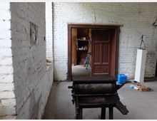 Ein rustikaler Innenraum mit weißen Ziegelwänden und einer braunen Holztür, die zu einem anderen Raum führt. Im Vordergrund steht eine alte Druck- oder Walzmaschine, während im Hintergrund ein Regal, ein Fahrrad und weitere Gegenstände sichtbar sind.