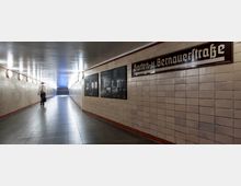 Unterführung mit beige gefliesten Wänden und runden Wandlampen, die gleichmäßig den Tunnel beleuchten. An der rechten Wand ist ein Schild mit der Aufschrift „Gaten-u. Bernauerstraße“ sowie Informationstafeln angebracht; im Hintergrund führen Treppen nach oben.