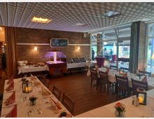 Ein festlich eingedeckter Speisesaal mit langen Tischen, geschmückt mit weißen Tischdecken, roten Servietten und kleinen Blumenarrangements. Im Hintergrund steht ein Buffetbereich, die Ziegelwand ist dekoriert mit Wandlampen und einem Gemälde, und durch große Fenster fällt Tageslicht herein.