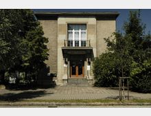 Fassade eines Kulturhauses in schlichtem Baustil mit einer zentralen Eingangstreppe und doppelflügeliger Holztür, umgeben von Bäumen und Grünfläche. Über der Tür befindet sich ein Schild mit der Aufschrift „Kulturhaus“.