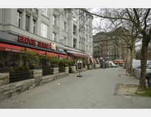 Straßenszene mit einem Restaurant („Block House“) in einem mehrstöckigen Gebäude mit heller Fassade. Vor dem Restaurant befinden sich eine Terrasse mit Pflanzen, Sitzbereichen und Schildern, daneben Gehweg, Bäume ohne Laub und geparkte Fahrräder.