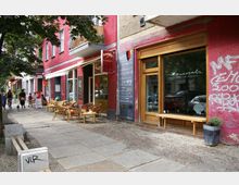 Straßenszene in einer Stadt mit Cafés und einem Restaurant in einem rosa Fassadengebäude. Vor den Geschäften stehen Tische und Stühle auf dem Gehweg, teils beschattet von Bäumen, und links im Hintergrund sind Passanten sowie eine Person mit Fahrrad zu sehen.