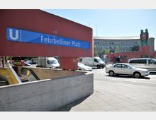 U-Bahnhof Fehrbelliner Platz in Berlin mit roter, moderner Architektur und einem blauen Stationsschild. Im Vordergrund befindet sich der Eingang mit Rolltreppen, im Hintergrund sind eine Straße mit Autos sowie ein großes Gebäude zu sehen.