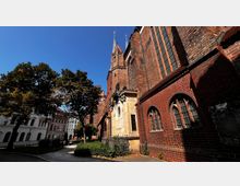 Backstein-Kirche mit gotischer Architektur, spitzbogigen Fenstern und einem dekorativen Eingangsportal. Im Vordergrund sind Bäume und ein gepflasterter Weg zu sehen, umgeben von weiteren historischen Gebäuden.