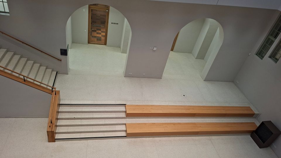 Innenansicht eines modernen Foyers mit hellen Steinböden, breiten Treppenstufen und Holzhandläufen sowie langen, niedrigen Holzpodesten. An der Rückwand befinden sich große Rundbögen und eine Holztür mit der Beschriftung „Museum“, daneben Fenster an der rechten Wand.