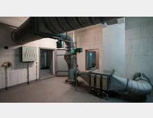 Ein industrieller Technikraum mit Lüftungsrohren und einer Schalttafel an einer Betonwand. Im Hintergrund sind schwere Metalltüren und angrenzende Räume sichtbar.