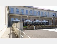 Zweistöckiges Gebäude mit heller Fassade, großen Fenstern und einem überdachten Außenbereich mit mehreren Sonnenschirmen. Im Vordergrund sind ein gepflasterter Weg, Geländer und eine schmale Rampe sichtbar, umgeben von Blumenbeeten und Laternen.