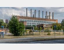 Backstein-Industriegebäude mit mehreren vertikalen Schornsteinen auf dem Dach, umgeben von Grünflächen und metallischen Rohrleitungen. Im Vordergrund sind ein Gehweg, eine Straße sowie ein Schild mit der Aufschrift „Werkstraße“ zu sehen.