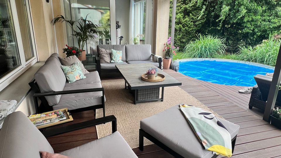Überdachte Holzterrasse an einem Wohnhaus mit Lounge-Sitzmöbeln, Couchtisch und Teppich, angrenzend an einen runden, blauen Pool. Im Hintergrund stehen hohe, dichte Hecken und Ziergräser am Rand des Gartens.