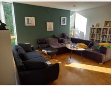 Wohnzimmer mit einer Sitzecke aus dunklen Sofas und einem schwarzen Ledersessel mit Fußhocker. Eine grüne Wand mit Bildern, ein Bücherregal an der weißen Wand sowie große Fenster, die Tageslicht hereinlassen, prägen den Raum.