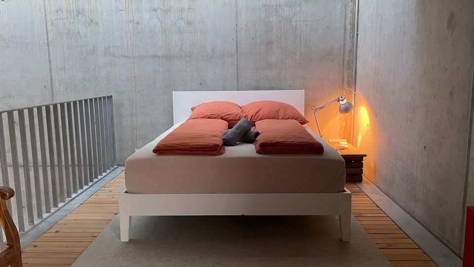 Minimalist bedroom with exposed concrete walls and a white platform bed dressed in rust-colored bedding, set on a rug over wood plank flooring. A small bedside table with an angled lamp sits to the right, and a metal railing lines the left side like a loft edge.