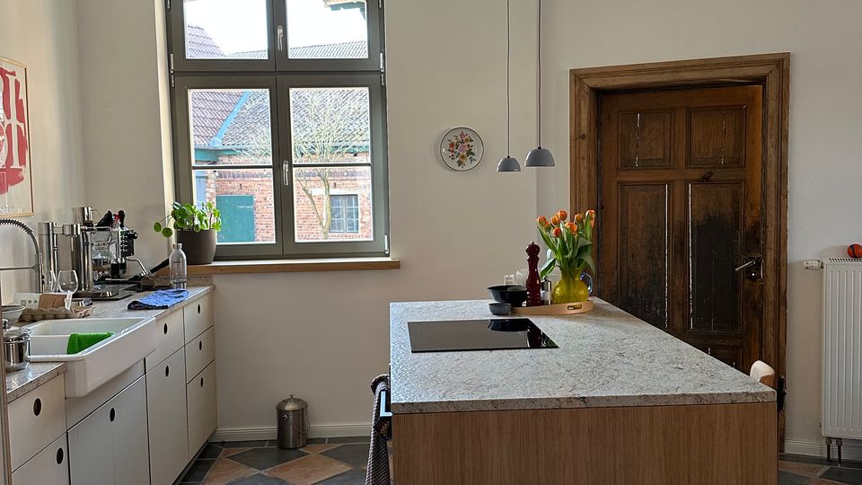 Helle Wohnküche mit großem Sprossenfenster, weißen Einbauschränken entlang der linken Wand und einer zentralen Kücheninsel aus Holz mit heller Arbeitsplatte und Kochfeld. Sichtbare Pendelleuchten an Kabeln, eine dunkle Holztür rechts und ein diagonal verlegter Fliesenboden in Blau- und Rosatönen.