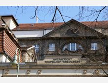 Fassade des Amtsgerichts Tempelhof-Kreuzberg mit Schmuckelementen wie Blumenreliefs und Kletterpflanzen. Das historische Gebäude besitzt rotes Ziegeldach, weiße Fenster sowie erkennbaren Schriftzug auf der Steinfront.