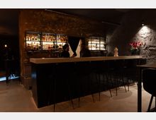 Eine moderne Bar mit einer langen weißen Theke und schwarzen Barhockern vor einer Wand aus unverputztem Ziegelstein. Im Hintergrund sind beleuchtete Regale mit Flaschen und Gläsern, sowie eine Vase mit roten Blumen als Dekoration sichtbar.