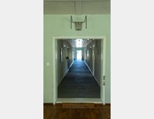 Ein langer, schmaler Flur mit Teppichboden, weißen Wänden und eingelassenen Deckenleuchten, der von einem Raum mit Parkettboden aus sichtbar ist. An den Wänden des Flurs hängen vereinzelt Bilder oder Tafeln, und am Ende des Ganges ist eine Lichtquelle oder ein Fenster erkennbar.