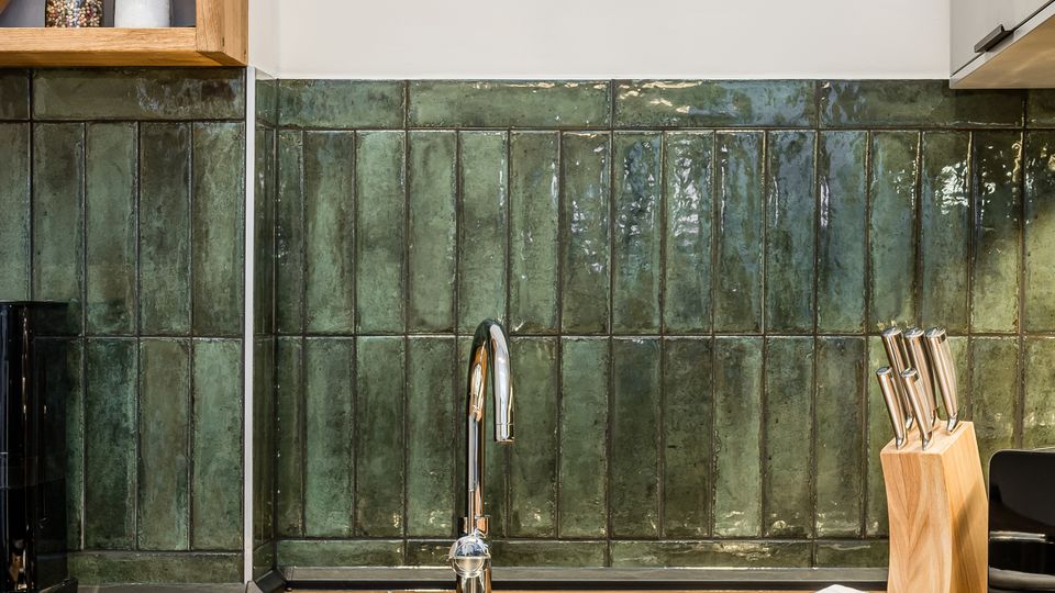 Moderne Küchenzeile mit Holzarbeitsplatte und Edelstahlspüle, darüber eine grün glasierte Fliesenrückwand. Links ein offenes Holzregal mit Gewürzstreuern, rechts ein Messerblock vor hellen Schränken.