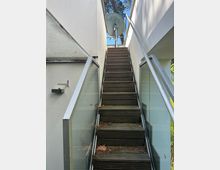 Außentreppe aus Holz zwischen zwei weißen Gebäudewänden, mit Glasgeländern und Metallhandlauf auf beiden Seiten. Oben führt die Treppe zu einer kleinen Plattform mit großer Satellitenschüssel vor blauem Himmel und Bäumen im Hintergrund.