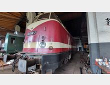 Eine rot-beige Diesellokomotive mit der Nummer "118 748-3" steht in einer Werkstatthalle mit hölzernem Dach und Ziegelwänden. Links ist ein grüner Waggon zu sehen, und verschiedene Werkzeuge sowie Maschinen stehen im Umfeld verteilt.