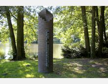 Eine zylindrische Steinsäule mit der Inschrift "KARL LIEBKNECHT" steht auf einer grünen Wiese, umgeben von hohen Bäumen. Im Hintergrund ist ein ruhiger See mit dicht bewachsenen Ufern zu sehen.