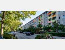 Mehrstöckiges Wohnhaus mit farbigen Balkonen in einer Wohnsiedlung, flankiert von einer schmalen Straße mit parkenden Autos und von großen, schattenspendenden Bäumen gesäumt. Die Umgebung wirkt grün und gepflegt mit Sträuchern und kleinen Gärten.