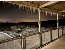 Blick von einer überdachten Terrasse mit Metallgeländer auf eine schneebedeckte Landschaft bei Nacht. Eiszapfen hängen vom Dach, im Hintergrund sind Büsche, ein verschneiter Gehweg und ein leuchtendes rotes Licht am Horizont sichtbar.