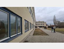 Langer, mehrstöckiger Gebäudekomplex mit hell verputzter Fassade und blau gerahmten Fenstern entlang eines gepflasterten Gehwegs. Rechts daneben liegen Rasenflächen mit kahlen Bäumen; im Hintergrund steht ein weiterer flacher Gebäudetrakt unter bedecktem Himmel.