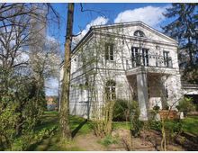 Weiße zweistöckige Villa mit klassizistischen Elementen, darunter ein Dreiecksgiebel und Balkon, umgeben von einem Garten mit blühenden Bäumen und Buschwerk; im Hintergrund blauer Himmel mit vereinzelten Wolken.