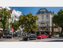Straßenszene in einer Stadt mit einem hellen, historischen Wohngebäude im Vordergrund, das verzierte Fassadendetails und große Fenster aufweist. Umgeben von Bäumen, parkenden Autos und Fahrradständern sowie einem Café im Erdgeschoss des Gebäudes.
