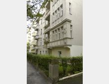 Mehrstöckiges Wohngebäude im Stil der Gründerzeit mit hellgrauer Fassade, Erkern und Ornamenten über den Fenstern. Vor dem Gebäude befindet sich ein kleiner, begrünter Vorgarten mit Hecken und einem Gehweg.