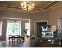 Ein helles Café mit hohen Decken, verziert mit Stuck und einem Kronleuchter, zeigt eine gemütliche Sitzecke mit Holztischen und -stühlen vor großen Fenstern. Rechts im Vordergrund steht eine Vitrine mit Pflanzen und einem Angebot an Speisen oder Getränken.