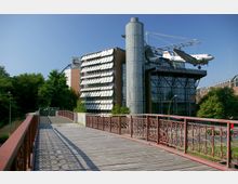 Holzbrücke mit Metallgeländer führt zu einem modernen Gebäude des Deutschen Technikmuseums in Berlin, an dem ein Flugzeug an einer Außenkonstruktion befestigt ist; umgeben von Bäumen und städtischer Umgebung.