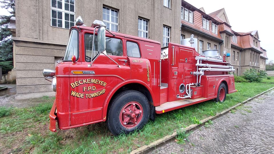 Rotes historisches Feuerwehrauto mit der Aufschrift „Beckemeyer F.P.D Wade Township“, geparkt auf einer Grünfläche vor einem beige-braunen Gebäude mit roten Ziegeldächern und Sprossenfenstern.