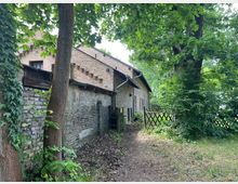 Backsteinbau mit Satteldach in ländlicher Umgebung, umgeben von Bäumen und Gebüsch. Ein kleiner Pfad führt zu einem Holztor neben einer verwitterten Backsteinmauer.