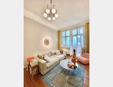 Wohnzimmer mit heller Einrichtung, bestehend aus einem beigen Sofa, einem runden Couchtisch und einer rosa Sitzliege. Große Fenster mit gelben Vorhängen lassen viel Licht ein, und an der Decke hängt eine moderne, runde Lampe; auf dem Holzboden liegt ein blauer Teppich.