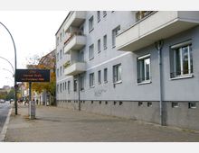 Mehrstöckiges Wohnhaus mit hellgrauer Fassade und Balkonen an einer Straßenecke. Auf dem Gehweg stehen eine digitale Verkehrsanzeige mit Stauhinweis sowie einige Fußgänger, im Hintergrund Bäume und weitere Gebäude.