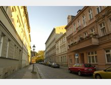 Eine gepflasterte Straße in einem historischen Wohnviertel mit mehrstöckigen Gebäuden im Stil des 19. Jahrhunderts auf beiden Seiten. Parkende Autos säumen die Straße, und Blumen schmücken die Balkone der Gebäude; eine Laterne mit einem Werbeplakat steht am Gehweg.