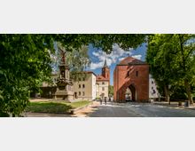 Historisches Stadttor aus rotem Backstein mit spitzbogigem Durchgang, umgeben von Bäumen und gepflasterten Wegen. Links steht ein Denkmal mit einer geflügelten Statue, und im Hintergrund ist eine Kirche mit einem spitzen Turm zu sehen.