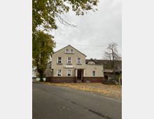 Zweistöckiges Wohnhaus mit heller Fassade und roten Klinkersteinen im unteren Bereich, gelegen an einer ruhigen Straße mit Herbstlaub auf dem Boden. Im Vordergrund stehen Bäume mit spärlichem Laub, die einen Teil des Himmels rahmen.