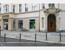 Fassade eines Eckgebäudes mit sandfarbener Steinverkleidung, großen Fenstern und einem hölzernen Eingangstor mit dekorativem Steinbogen. Links befindet sich eine Apotheke mit Schaufenstern, Fahrrädern davor und gepflastertem Gehweg.