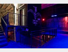 Dunkler Innenraum eines Nachtclubs oder Veranstaltungsortes mit Treppengeländern und bunt beleuchteten Wänden. Neon-Schriftzüge wie „ACCEPT THE FUTURE TRUE SPIRIT“ sind an einer schwarzen Wand sichtbar, neben einer Zeichnung eines stilisierten Vogels.