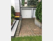 Einfahrt zu einer Garage mit gepflastertem Weg, umgeben von Zierpflanzen und Hecken. Links führen Stufen aus Stein nach oben, und rechts befindet sich die Garage mit einem weißen Tor sowie einer zusätzlichen grauen Tür.