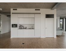 Kompakte, moderne Kücheneinheit in einem offenen, minimalistischen Raum mit weißem Schrankdesign, integrierter Kaffeemaschine und Holzfußboden. Im Hintergrund sind Betonwände, ein angrenzendes Sofa und große Fenster zu erkennen.