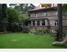 Zweistöckiges Wohnhaus mit einer Fassade aus Putz und rotem Backstein, umgeben von einem Garten mit Rasenfläche, Bäumen und Blumenbeeten. Im Vordergrund befindet sich eine Liege, und quer über den Garten spannen sich Lichterketten.