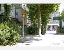 Eingang zu einem Innenhof mit Bäumen und einem grünen Zaun. Im Hintergrund sind ein Gebäude mit großen Fenstern sowie ein Außenbereich mit Sitzmöbeln und Menschen zu sehen.