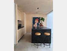 Moderne Küchenzeile mit Holzverkleidung und Betonboden, zentral ein schwarzer Küchenblock mit einer Pflanze und zwei Hockern mit hellbraunen Polstern. An der Rückwand hängt ein buntes Kunstwerk.